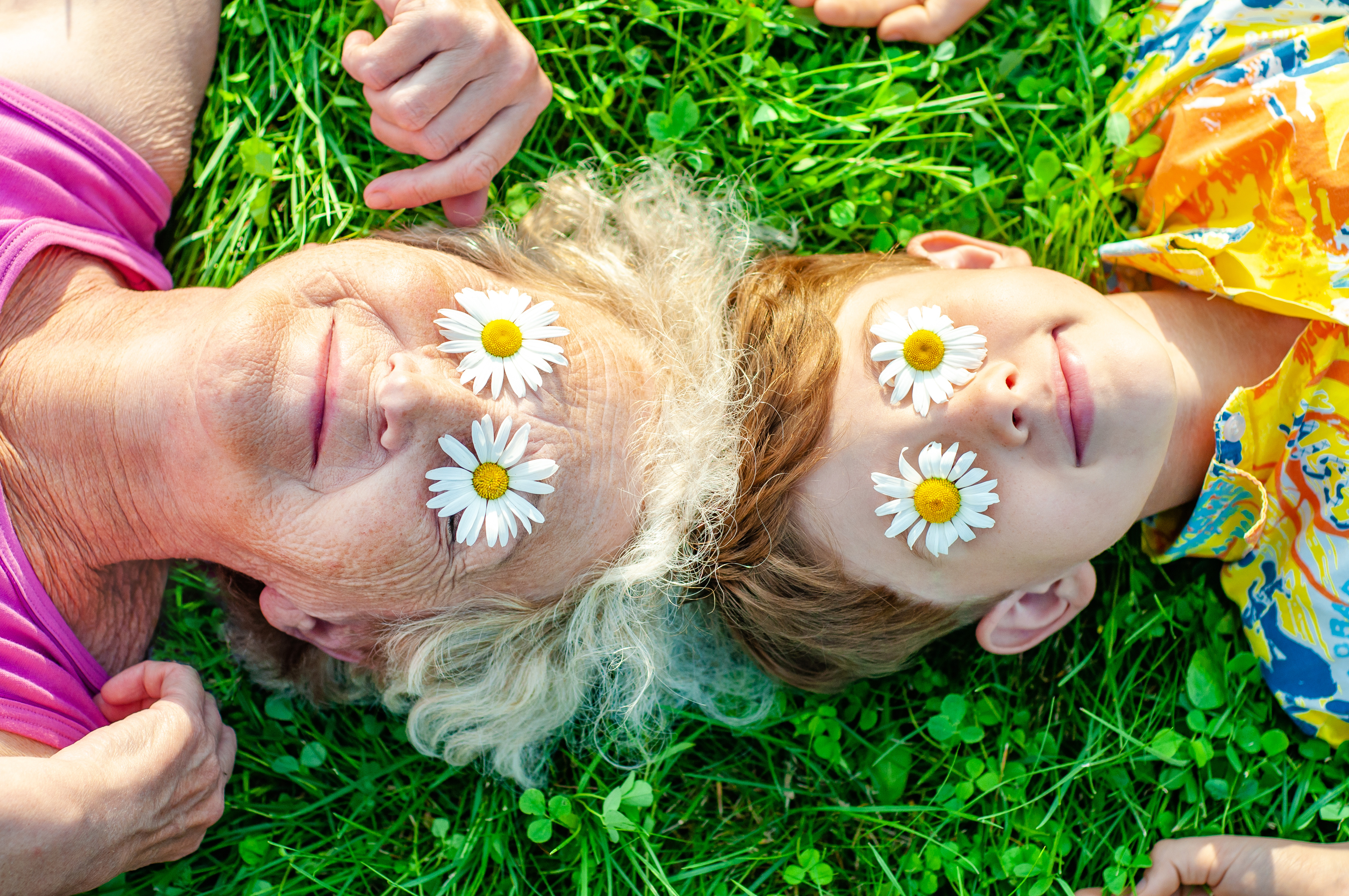 Happy family - grandmother with grandson having fun lying on the lawn with daisies on their eyes. Summer vacation with my grandmother. Family relationships and friendship